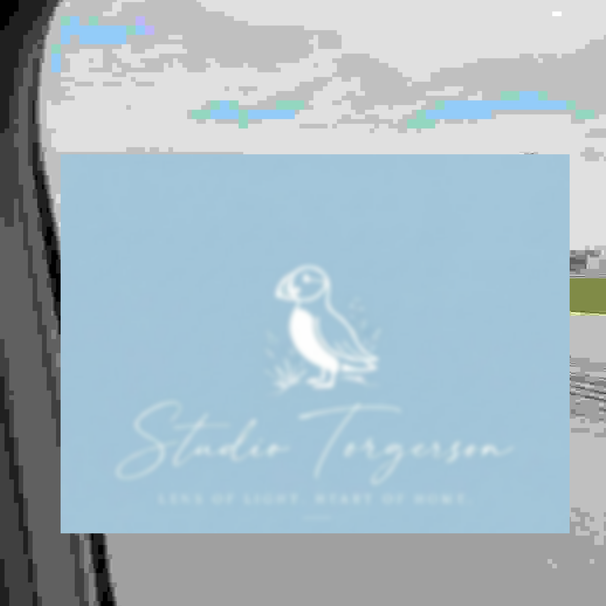 A square light blue card or label featuring a white puffin illustration and the text 'Studio Torgerson' with smaller text below, shown against a gray background with blurred blue elements.