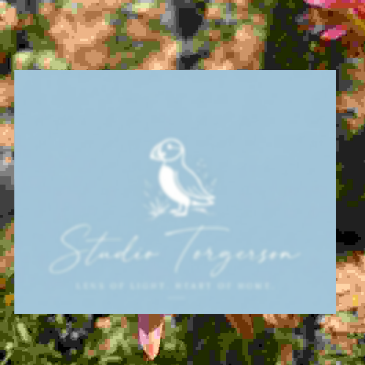 A square light blue card or label with a white puffin illustration and the text 'Studio Torgerson' and 'Lark of Light, Heart of Home,' centered over a blurred floral background.