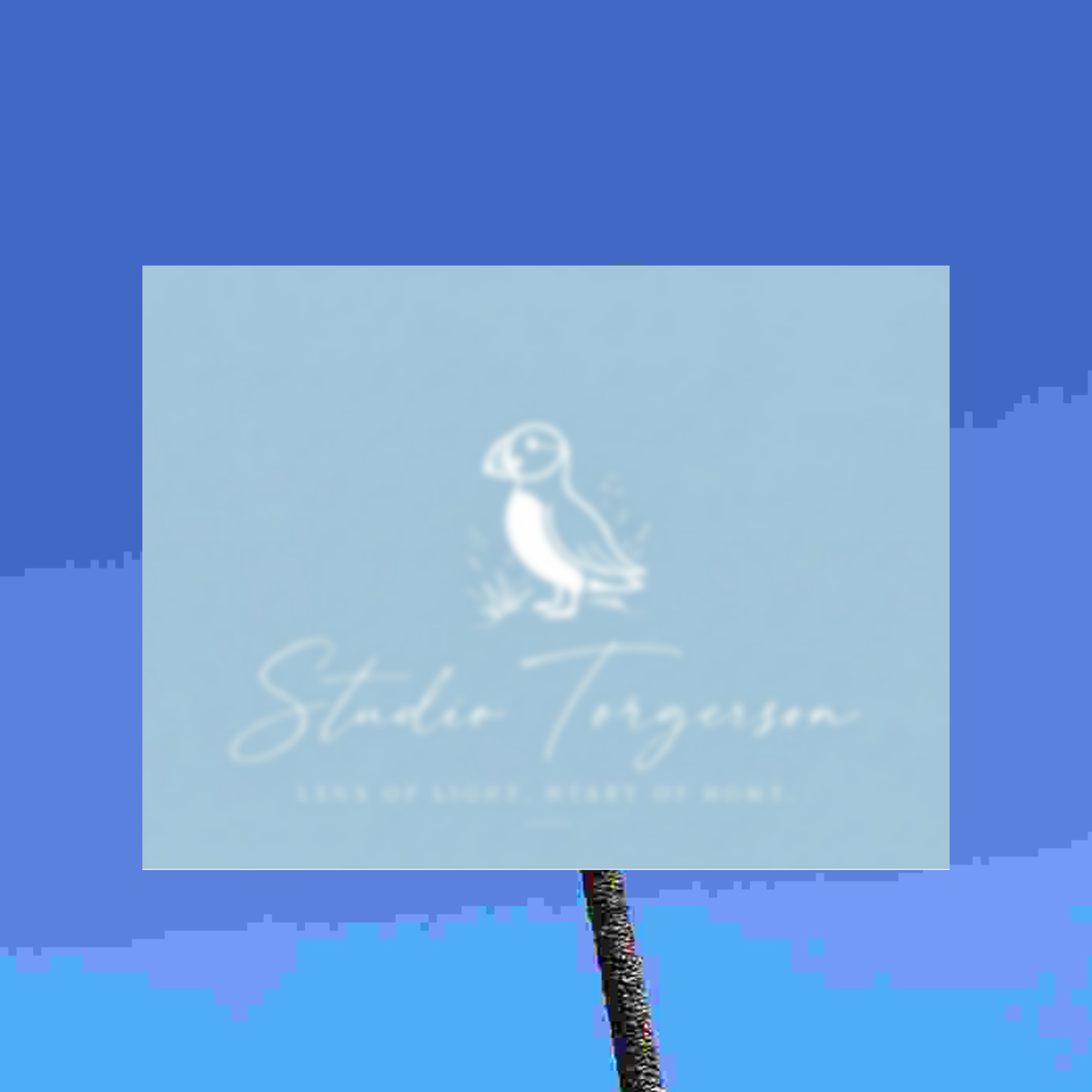 A square light blue card or print with a white puffin illustration and the text 'Studio Torgerson' and 'Lens of Light, Heart of Maine' on a blue background, mounted on a dark textured pole.