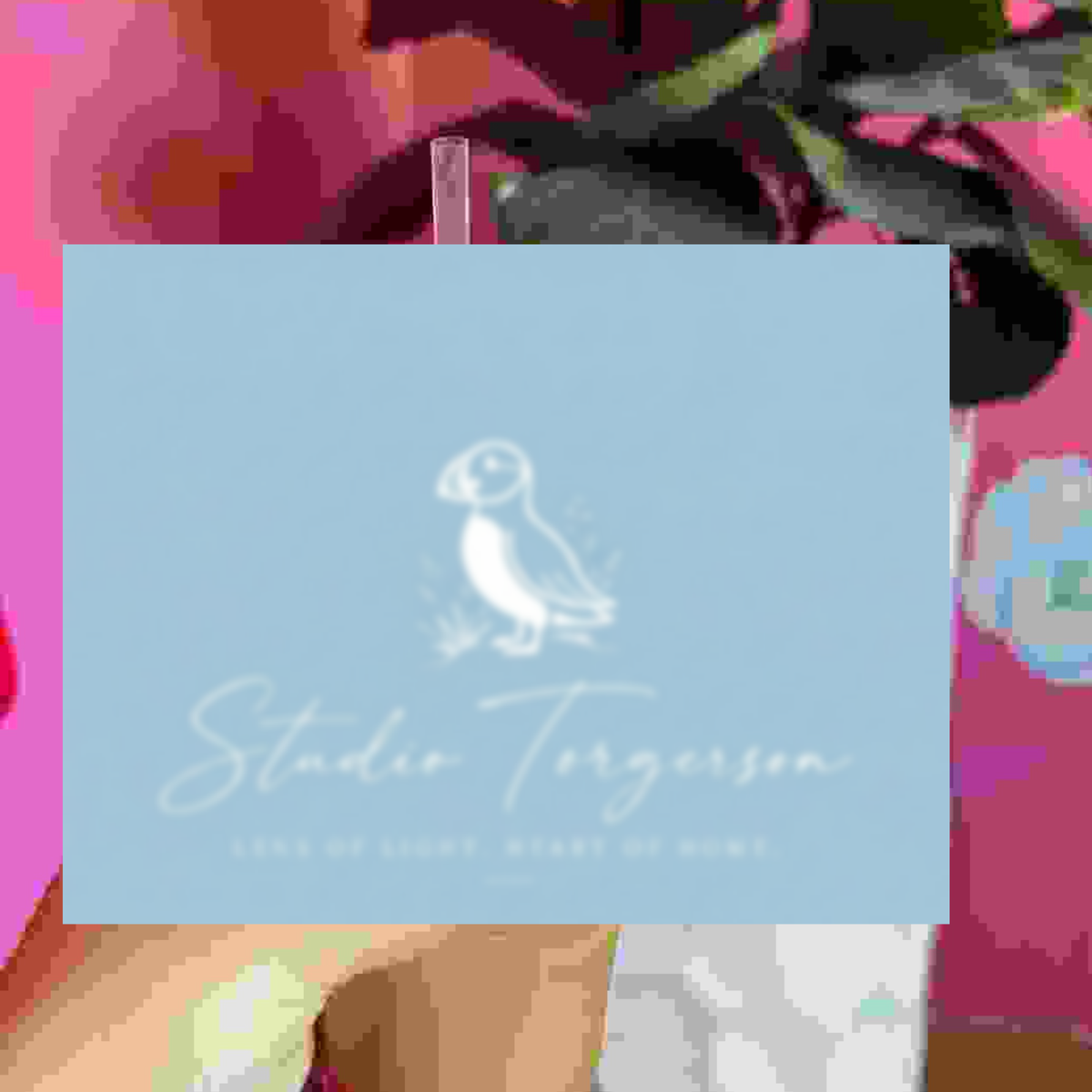 A square light blue card or print featuring a white puffin illustration and the text 'Studio Torgerson' with smaller text below, held against a colorful blurred floral background.