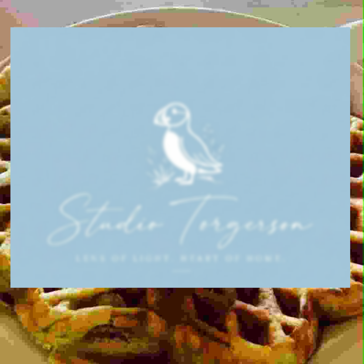 A square light blue card or label with a white puffin illustration and the text 'Studio Torgerson' and 'LONg OF LIGHT, HEART OF HOME' overlaid on a blurred background of waffles on a plate.